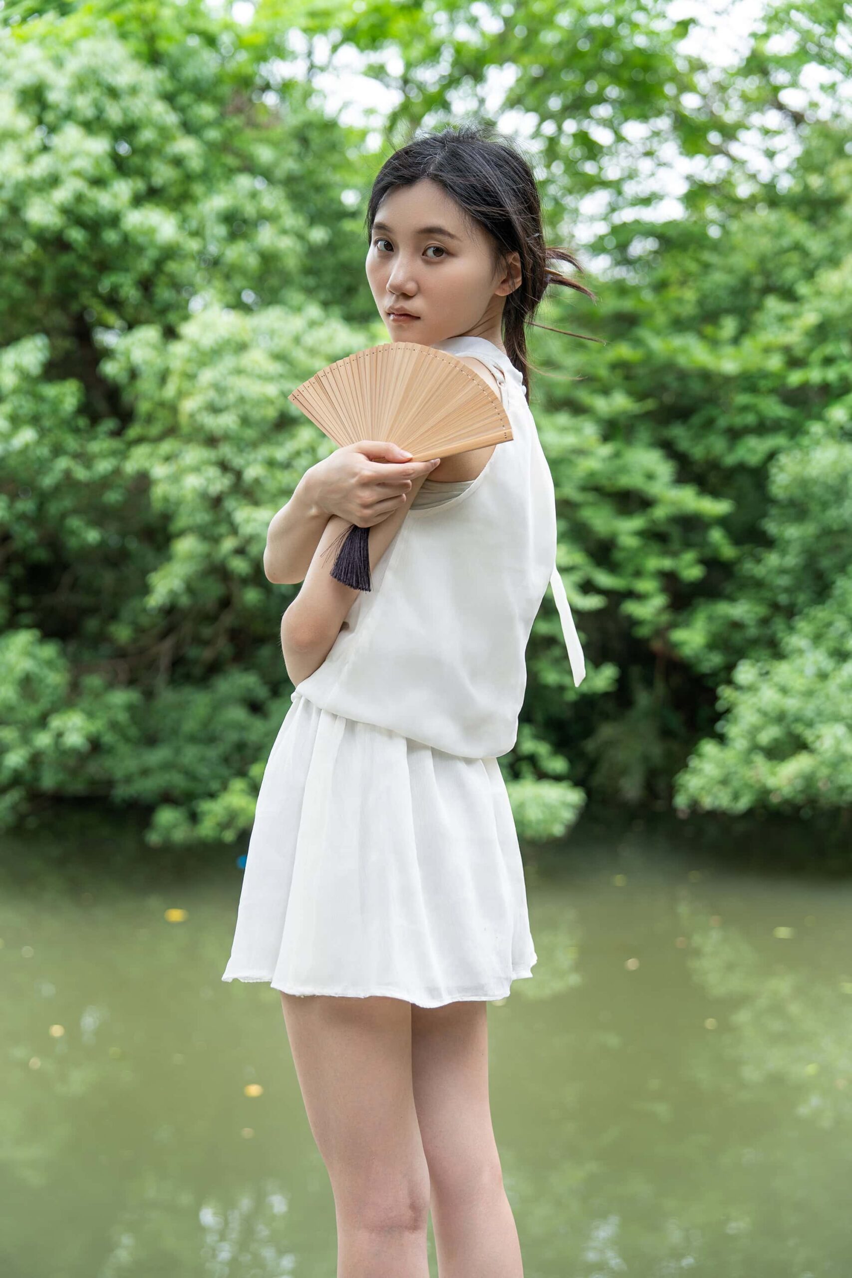 A woman in a white dress holding a Chinese hand fan, standing beside the calm waters of Xixi Wetland