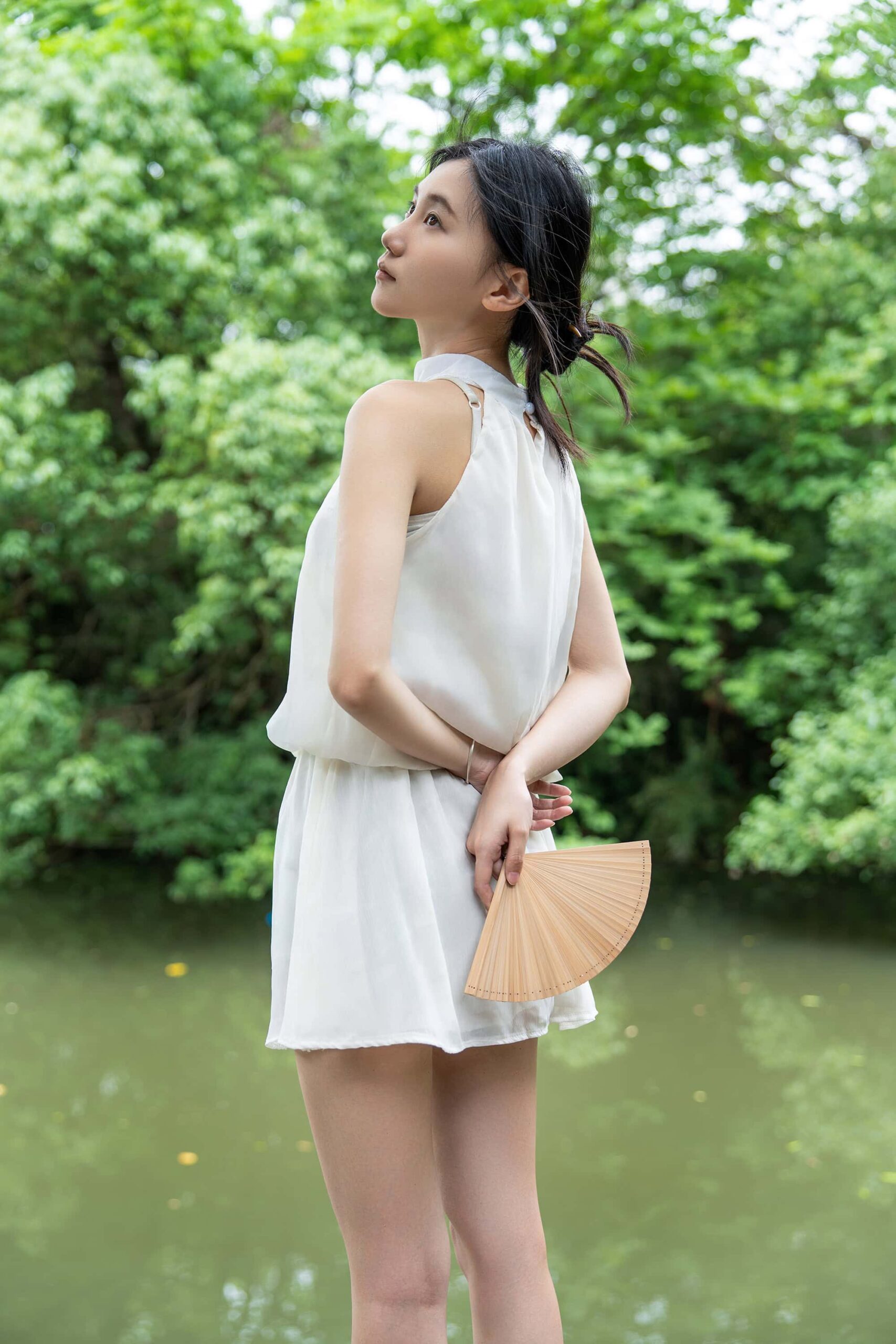 A woman stands gazing upwards with a serene expression, surrounded by nature at Hangzhou Xixi Wetland Park, wearing a minimalist white dress