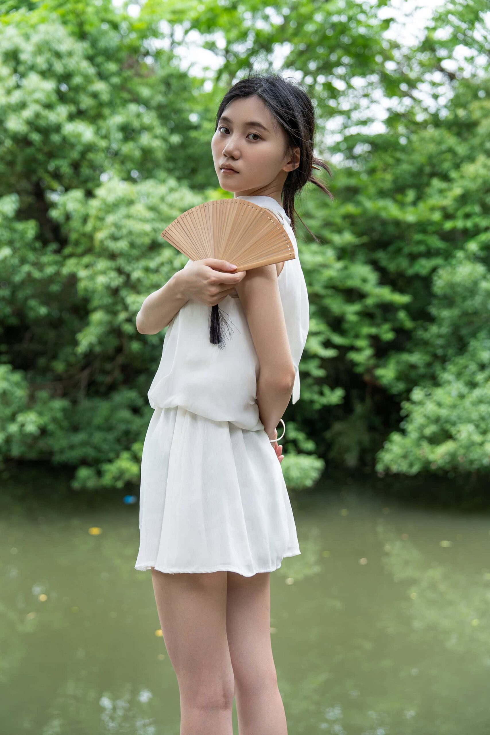 A young woman holding a traditional Chinese folding fan while standing gracefully in front of the serene Xixi Wetland