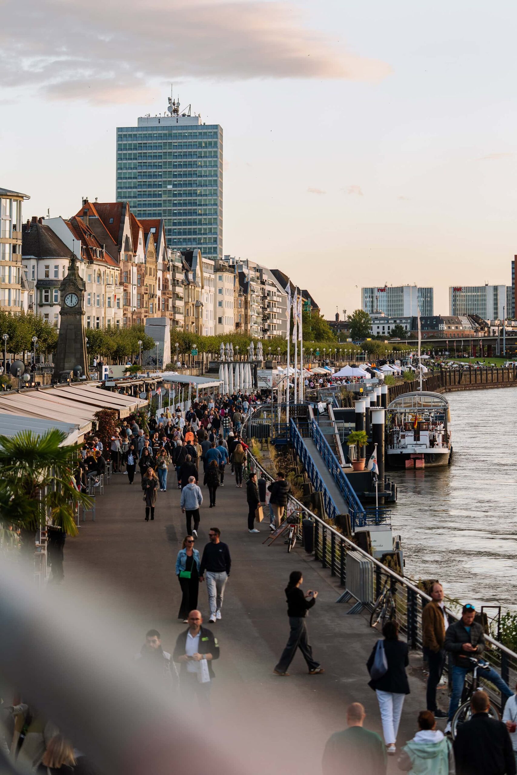 Evening scene on the Rhine embankment promenade in Düsseldorf, where cafés, riverboats, and historic facades line the lively walkway along the river