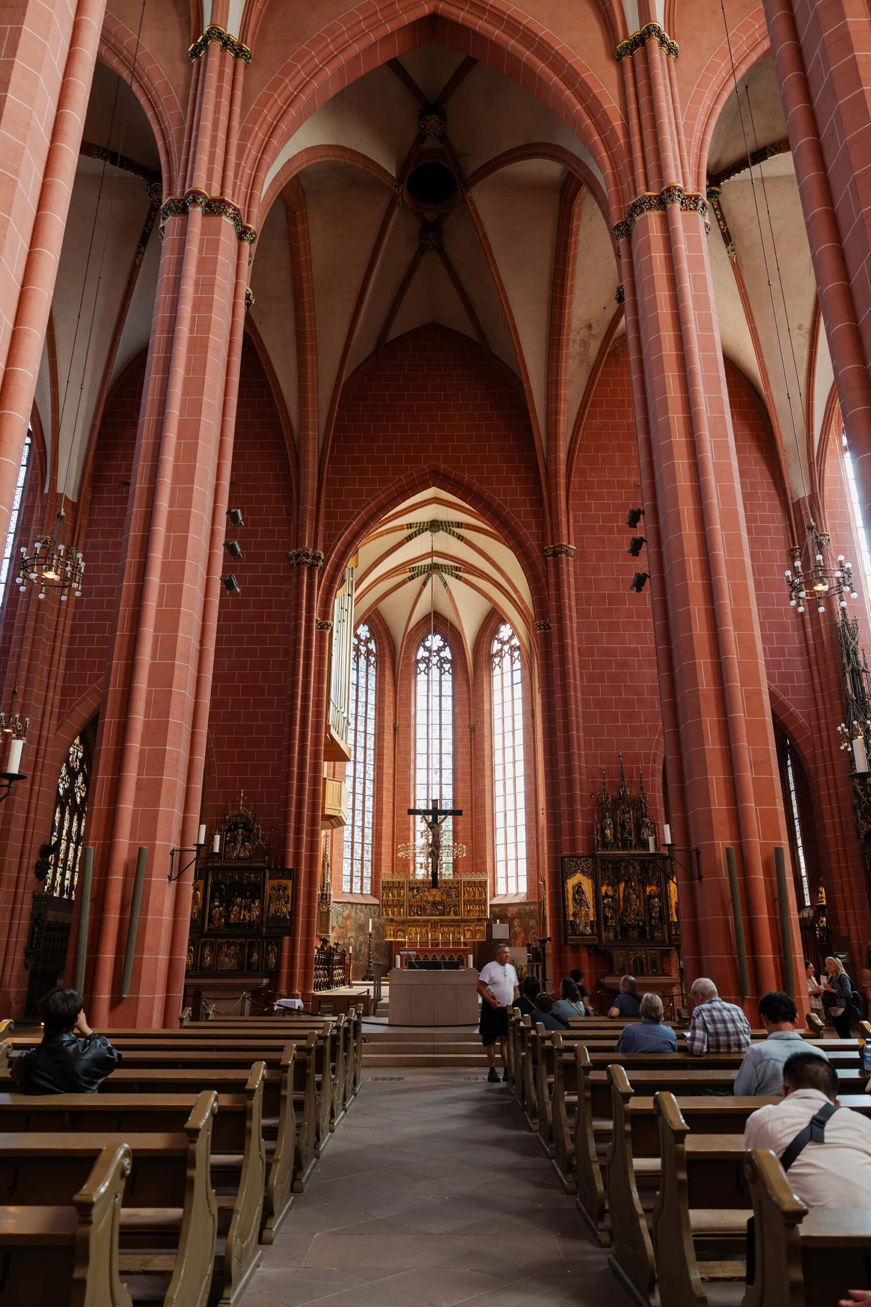 Majestic interior of the Frankfurt Cathedral (Kaiserdom Frankfurt), featuring towering red‑sandstone arches, the illuminated high altar, and visitors admiring this historic Gothic landmark