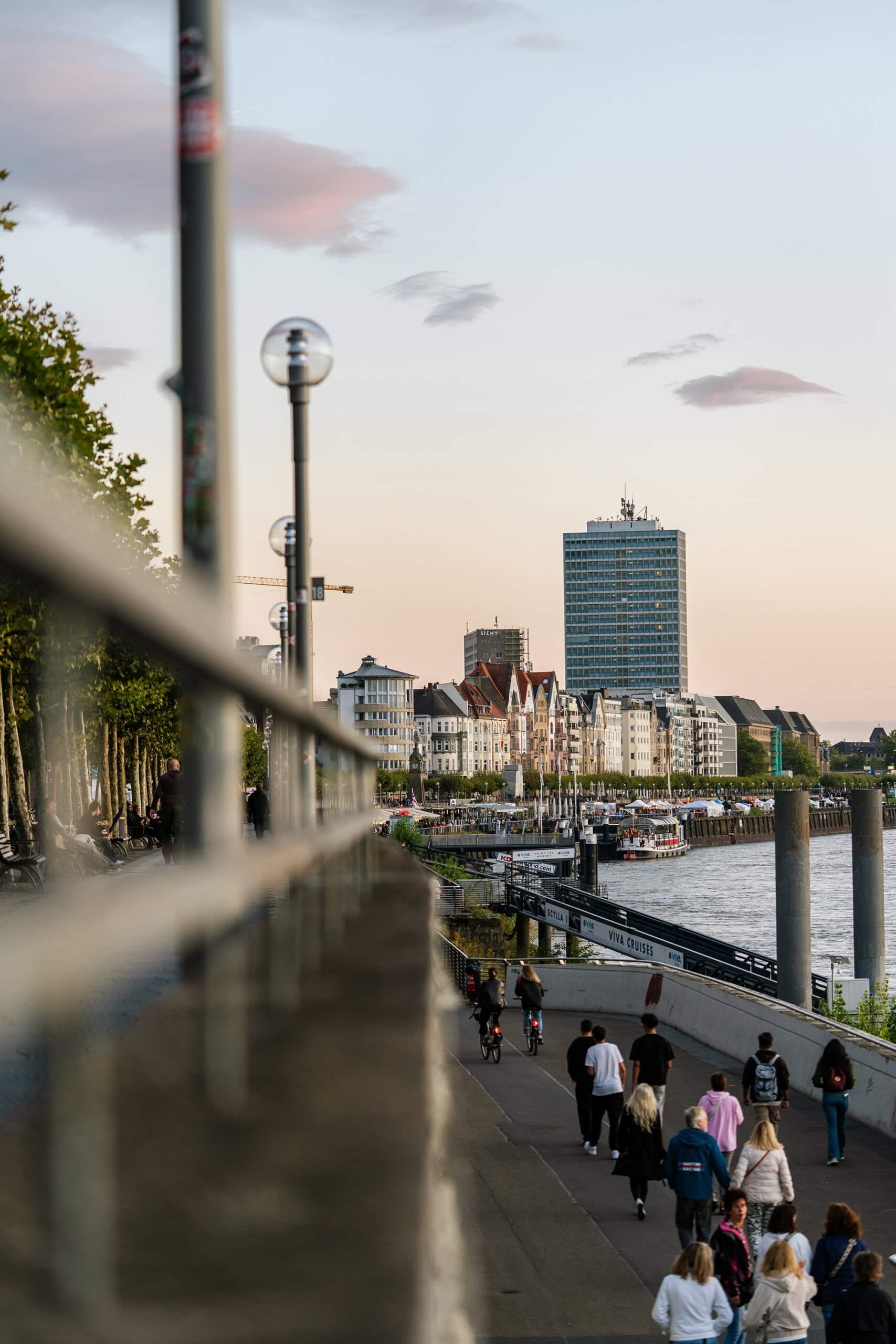 People strolling along the Düsseldorfer Rheinuferpromenade at sunset, with reflections of the sky on the Rhine River