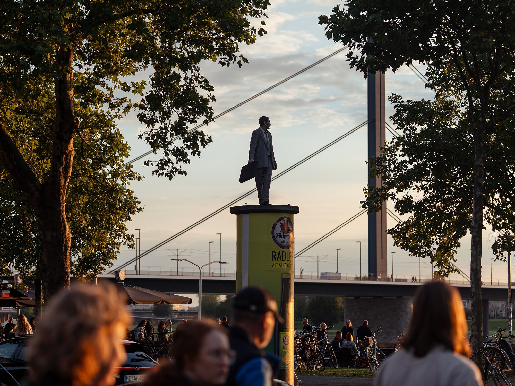 Statue near the Oberkasseler Bridge by the Tonhalle Düsseldorf, overlooking visitors gathered around Ehrenhof at dusk