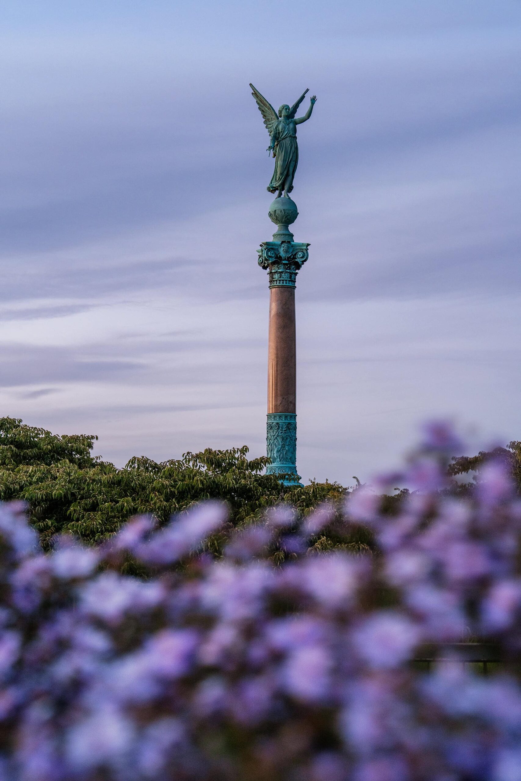 The Ivar Huitfeldt Memorial Column in Copenhagen, featuring the Roman goddess of victory standing atop the tall column, seen through soft-focus purple flowers