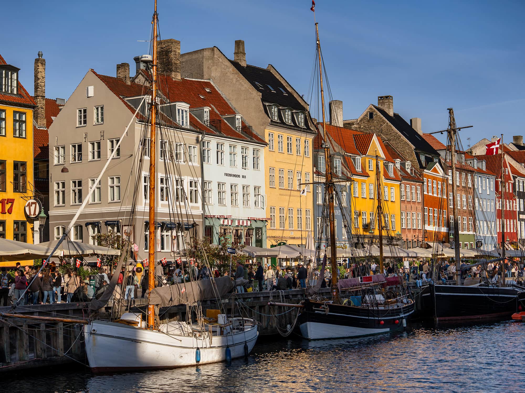 Boats moored along Nyhavn in Copenhagen, with rows of colorful historic houses and crowds enjoying the sunny harbor atmosphere