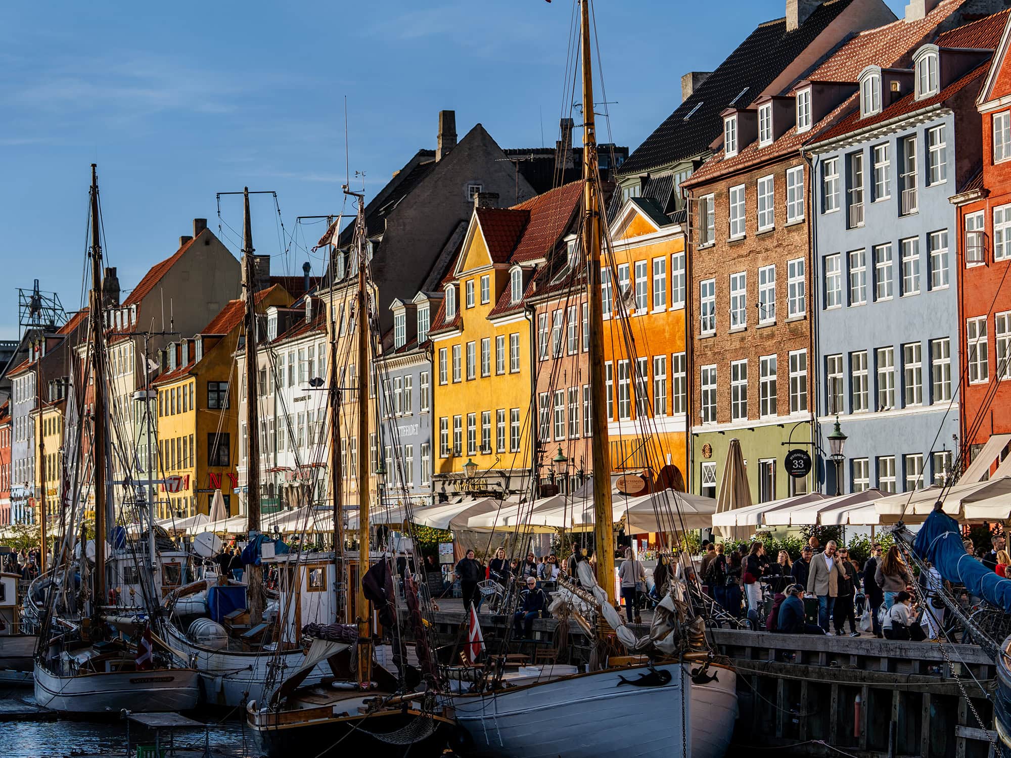 Sunlit Nyhavn colorful houses overlooking the Copenhagen canal, with moored boats and visitors strolling along the historic quay