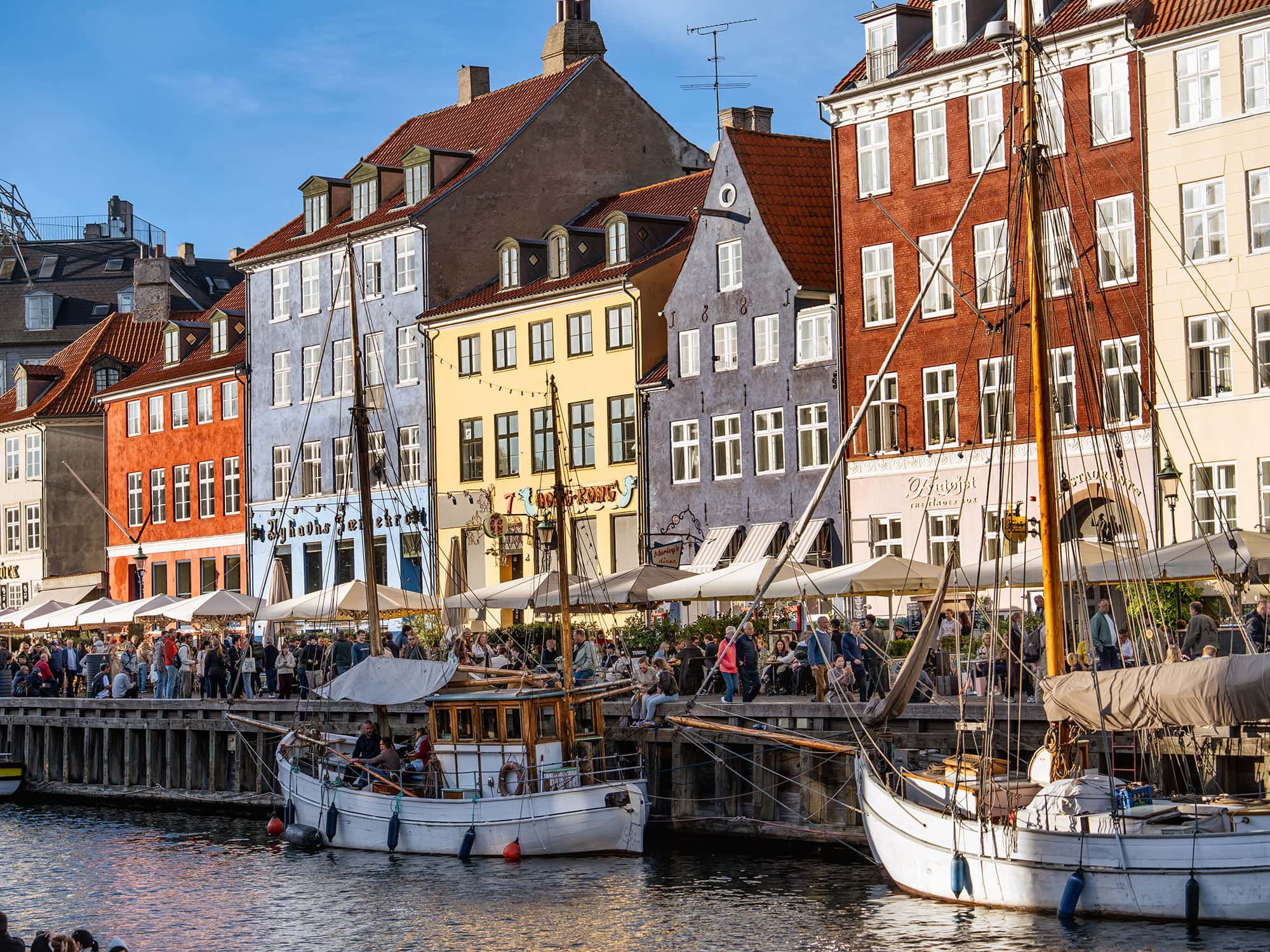 View of Nyhavn harbor in Copenhagen with classic wooden sailing boats, lively crowds, and bright Scandinavian houses lining the canal