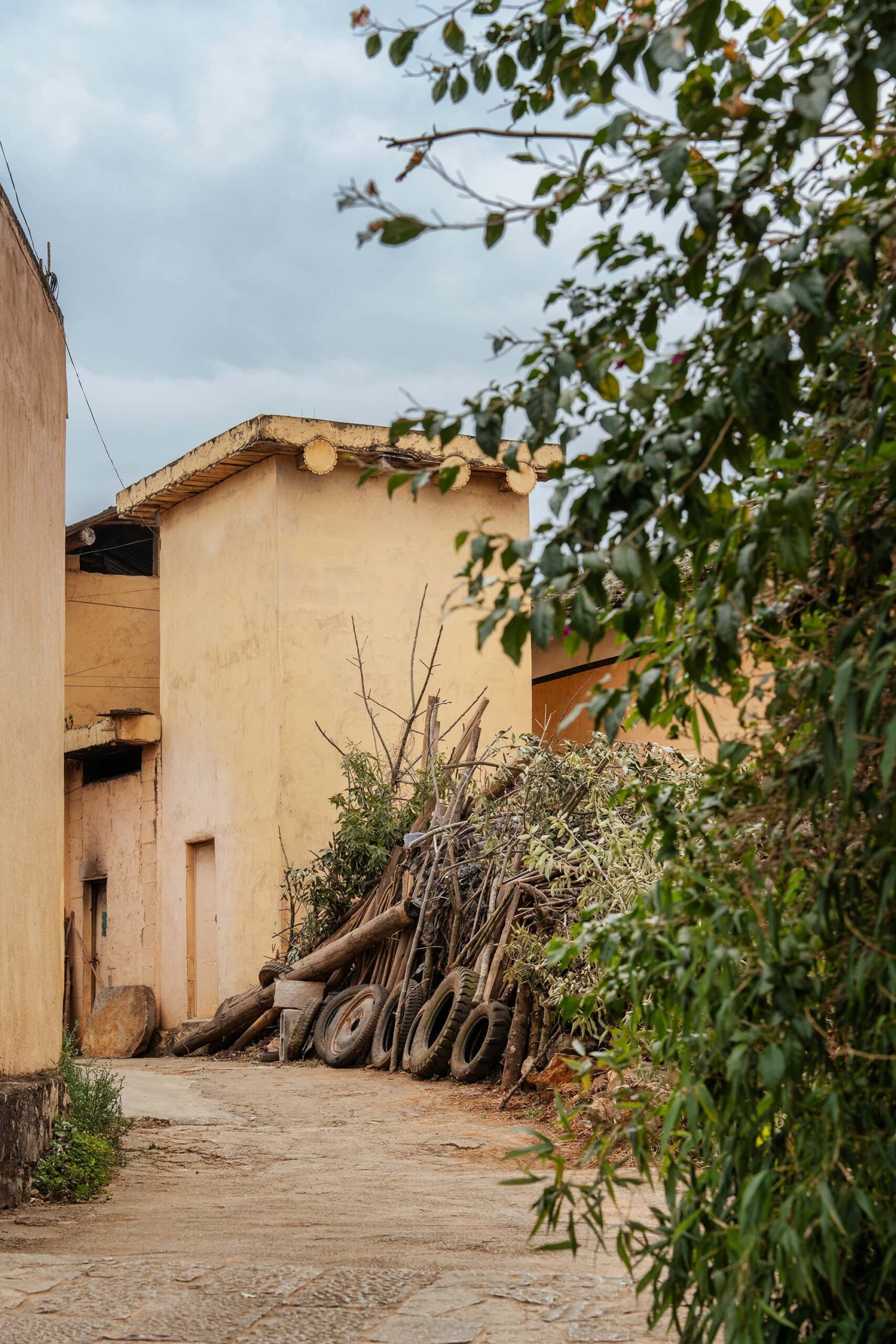 A narrow lane in Keyi Village with yellow earthen walls; a pile of firewood and several old tires stacked along the side, with leafy greenery in the foreground