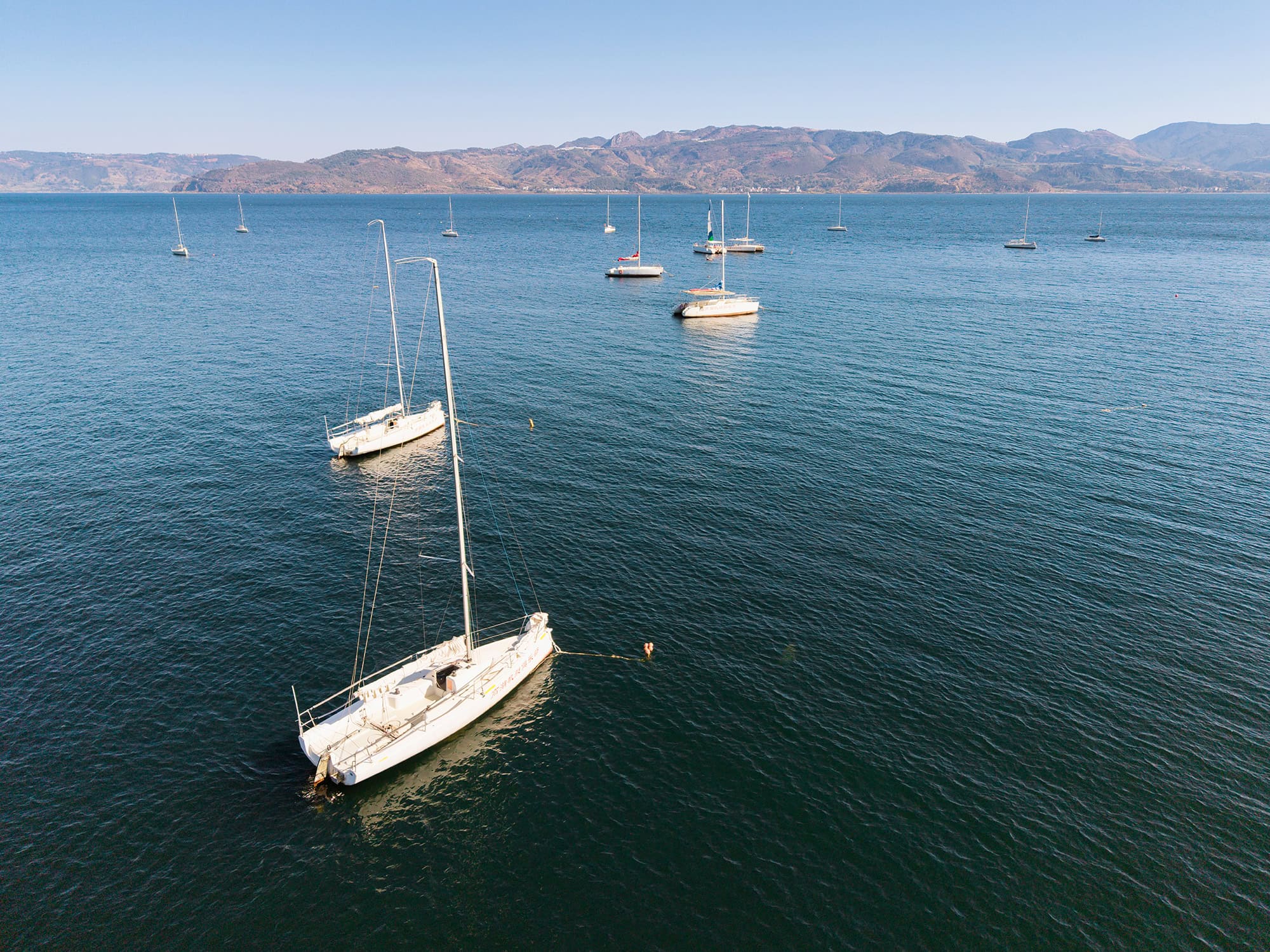 Aerial view of sailboats scattered across Fuxian Lake at Mingxing Fish Cave under a clear blue sky