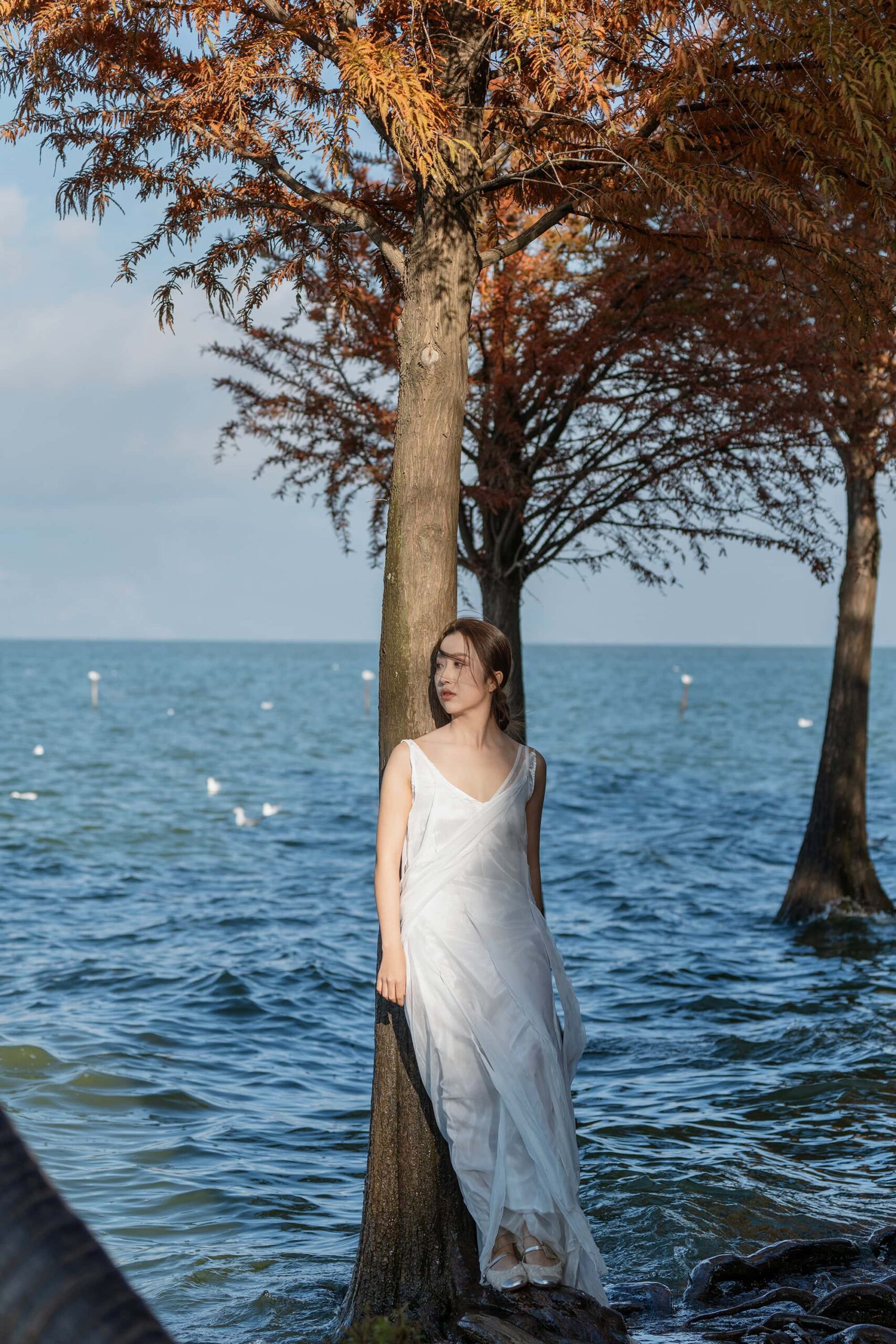 Full-body view of a woman in a white dress standing by a tree at Dianchi Lake