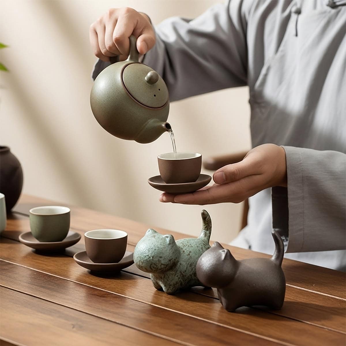 Person pouring tea into cup beside bronze and brown cat tea pets made of coarse clay with kiln-glaze texture