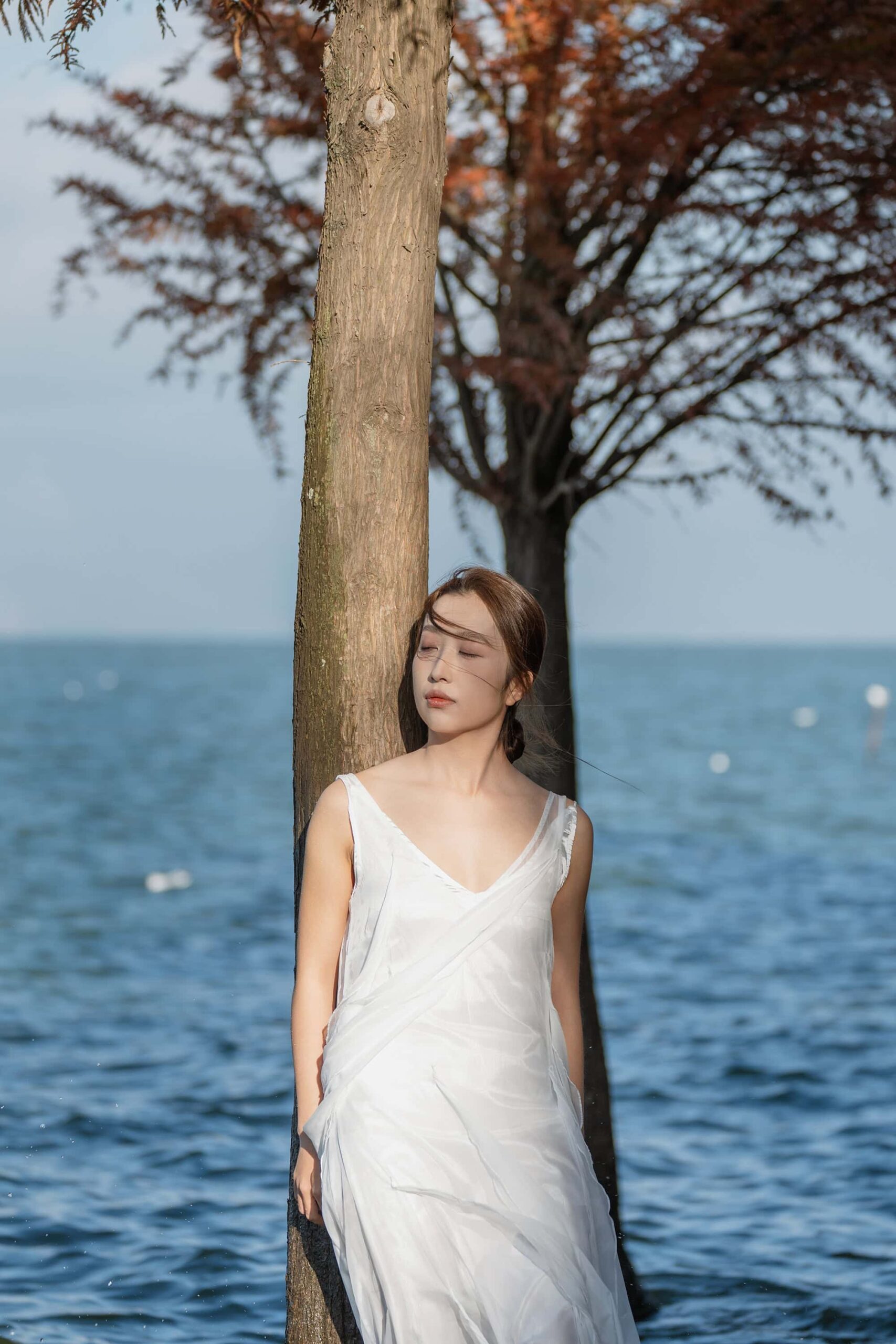 Woman in a white dress leaning against a lakeside tree at Dianchi Lake, with shallow blue water and distant gulls in the background