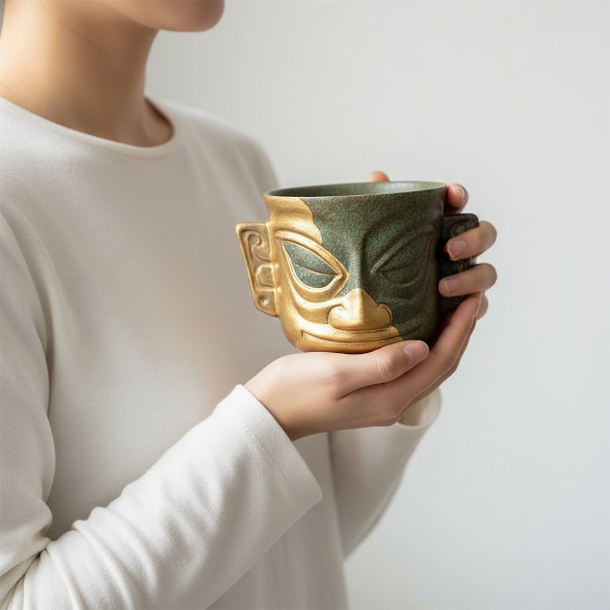 Handheld Chinese porcelain tea cup with a Sanxingdui civilization mask motif in gold and patina green