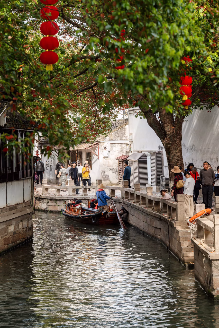 Boat with tourists gliding through a narrow canal lined with red lanterns and whitewashed houses in Tongli Water Town Suzhou​